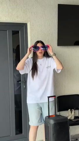 A young woman with long dark hair, wearing an oversized white t-shirt and light blue denim shorts, stands in front of a textured wall, holding large pink sunglasses up to her face. She is positioned next to a dark gray door on the left and a black speaker with an orange handle on the right. A black chair and a white bag are also visible to the right. The woman moves her arms and hands in a dancing or expressive manner, often raising her arms and gesturing. She briefly removes the sunglasses and then puts them back on. The lighting suggests an outdoor or well-lit indoor setting.