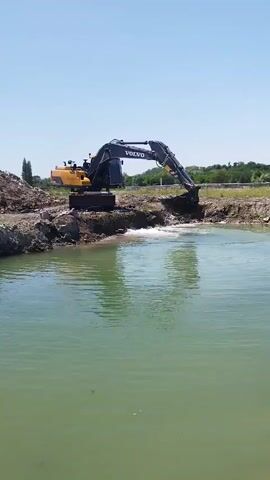 An excavator with the Volvo logo is digging in a body of water. The excavator is dark blue and yellow and is positioned on the bank of the water. It is lifting its arm, which has a bucket filled with water and dirt, out of the water. The water is green and calm, and the sky is clear and blue. There is a pile of dirt to the left of the excavator and some trees and vegetation in the background. The scene appears to be a construction site or a landscaping project.