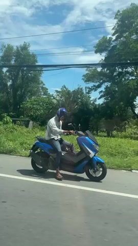 A man wearing a blue helmet, light gray hoodie, jeans, and sandals is riding a blue scooter on a paved road. He appears to be balancing himself by using his right foot to maintain his balance. The scenery consists of trees and greenery along the roadside, with power lines visible in the background. The weather appears to be sunny. The camera is positioned to the side of the scooter, moving at a similar pace, capturing the rider in motion.