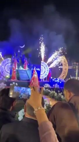 A wedding scene unfolds at night, with a bride center stage, surrounded by dancers. She stands in a red lehenga, complemented by traditional jewelry, while the dancers are also in red. The stage is bathed in blue lighting, decorated with white roses, and framed by semicircular and crescent-shaped designs. Pyrotechnics erupt on either side of the bride, adding to the spectacle. The scene is observed by an audience, some of whom are recording the performance with their phones.