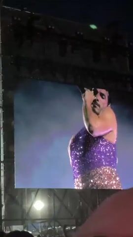 The video shows a person performing on a large screen at an outdoor concert. The performer is wearing a sparkly outfit, and the lighting is bright. The camera view is from within the crowd, looking up at the screen. The performer is shown dancing, singing into a microphone, and interacting with the audience.