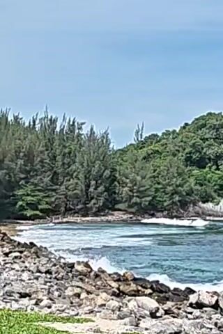 The video shows a rocky coastline with the ocean and a small, hilly island in the distance. The ocean is blue with some white waves breaking against the shore. The coastline is made up of rocks of various sizes. There are some trees and green vegetation near the rocks and on the island. The sky is light blue.