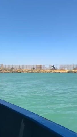 The video captures a view from a moving boat, with a blue and grey hull visible in the foreground, across turquoise-colored water towards a low-lying coastline. The sky is clear and bright blue, indicating a sunny day.

On the distant landmass, extensive industrial infrastructure is visible. Initially, a long, light-colored barrier or fence-like structure is seen, behind which some machinery operates. As the boat moves, a large-scale industrial conveyor belt system comes into view, extending from the shore into the water. This system, supported by multiple metal structures, appears designed for loading or unloading bulk materials.

At the end of the conveyor system, a large red bulk carrier ship is docked. The ship has a white superstructure and a dark blue logo with the text "zeunergy" clearly visible on its side. The conveyor system is actively engaged with the ship, suggesting ongoing loading or unloading operations. The scene depicts a busy industrial port facility, set against a backdrop of clear skies and vibrant waters. The location context provided is Unknown, Colombia.