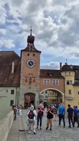 The video shows a street scene in a European town, likely Germany given the architecture. A prominent clock tower is visible, with a large clock face showing the time approximately at 3:10. Several people are walking on a cobblestone path in front of the tower and adjacent buildings. The buildings have a historical appearance, with tiled roofs and multi-story facades. Some people are looking at their phones, while others are talking or walking. The sky is partly cloudy. The atmosphere appears to be that of a tourist area.