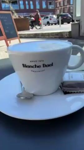 A cup of coffee sits on a saucer on a small round table outside a cafe. A small spoon and a packet of chocolate rest on the saucer. The cup is white and has the name "Blanche Dael" printed on it. The background shows a brick-paved outdoor area with a building across the way and a street with parked cars. A chalkboard menu stands next to the table listing the cafe's offerings. The scene is sunny. The camera slowly moves around the table, showing the surroundings.