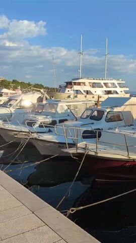 This video shows several boats docked in a harbor under a bright, partly cloudy sky. The boats are of varying sizes, with a large white yacht in the background and several smaller motorboats in the foreground. The smaller boats are white with red or dark hulls and are tied to a concrete dock. The water is dark and reflective, showing the shapes of the boats. The sun is casting long shadows, suggesting it is either early morning or late afternoon. The scene appears peaceful and serene.