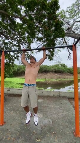 The video shows a man exercising on a pull-up bar at a park. The man, wearing a blue baseball cap, khaki shorts, and white socks with sneakers, grabs the bar with an overhand grip. He starts doing a modified version of a pull-up where he twists his body around the bar until he's hanging upside down with his feet in the air. The park is a concrete platform with orange metal supports for the pull-up bar frame. There is a green, overgrown pond behind the man, and trees are visible in the background. The day seems overcast.