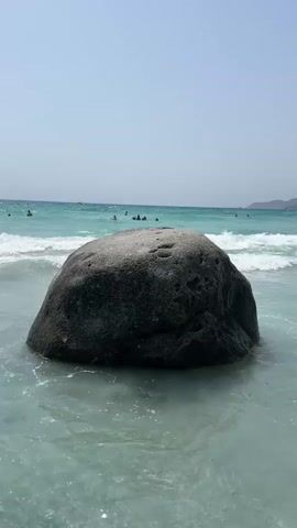 A large, dark boulder is partially submerged in clear turquoise water at the edge of a beach. Waves are gently breaking around the boulder, creating white foam. In the background, the ocean stretches to the horizon under a clear, pale blue sky. Several people are visible in the water, swimming and enjoying the ocean. The overall scene depicts a serene beach environment on a sunny day.