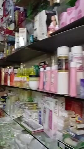 The camera pans across the interior of a small shop stocked with a wide variety of items. Shelves are densely packed with beauty products, containers, and other small household items. There are also decorative items like colorful pom-poms and flower arrangements. The shop appears somewhat cluttered with items arranged haphazardly on shelves and surfaces. The lighting is fluorescent and the overall environment suggests a small, family-run business. The front of the shop is visible through a metal gate that is partly rolled down.