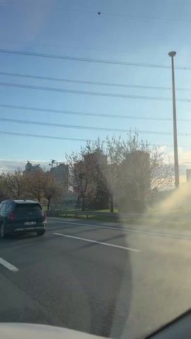 The video shows a car driving on a highway. On the side of the road there are buildings and trees. The sky is clear and blue. There is a sign that says "100 m." The sun is shining brightly, causing glare on the windshield. Other cars are also driving on the highway.