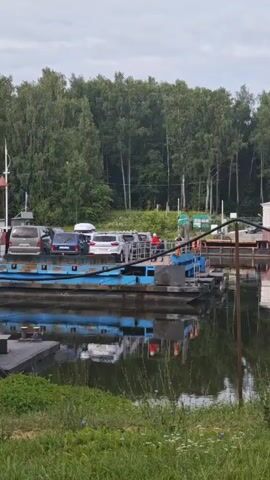 A ferryboat is loaded with several cars and people. The ferryboat is blue and sits on the water. Trees are visible in the background, along with a building that has peeling white paint and a small window. The sky is cloudy. A street light extends from the building, with wires attached to it. There is also a small structure with a pitched roof and a pink door. In the distance, there is a larger building with columns.