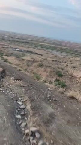 A first-person perspective video shows a bumpy dirt road or path as a vehicle moves along it. The road is uneven and appears to have some erosion. To the left of the road, there is a ditch with rocks. The surrounding environment is dry and appears to be a rural or undeveloped area with sparse vegetation and some distant structures or fences. The sky is visible in the background, appearing overcast or hazy. A side mirror of the vehicle is partially visible in the lower left corner of the frame in some shots. The video captures the motion of the vehicle and the roughness of the terrain.
