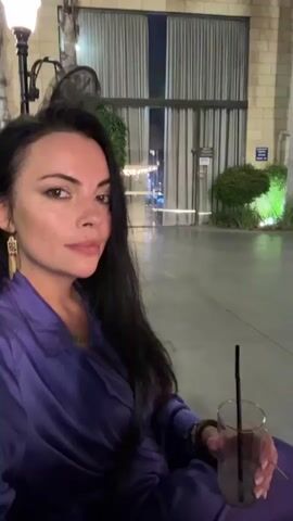 A woman with long, dark hair and heavy eye makeup is posing for a selfie in an outdoor seating area. She is wearing a shiny, loose-fitting purple top. In her right hand, she holds a glass with a clear liquid and a black straw. She is seated in a dark chair, and her posture suggests she is relaxed.

The background reveals a modern architectural setting with large glass doors and tall, thin curtains. Beyond the doors, soft lighting suggests it is evening or night. To the right of the building, palm trees are visible, illuminated by spotlights, casting a greenish glow on the surrounding bushes. The ground is a smooth, grey surface, likely a patio or walkway. In the distance, on an upper level of the building, several people can be seen standing and observing the area.

The overall mood is one of leisure and perhaps a sophisticated evening outing. The lighting and the woman's expression convey a sense of calm and self-assurance. There is a sign visible on the building in the background with some text, but it is too blurry to read. The scene appears to be a commercial or hospitality establishment, possibly a restaurant or bar with outdoor seating. The presence of palm trees and the architecture suggest a warm climate.