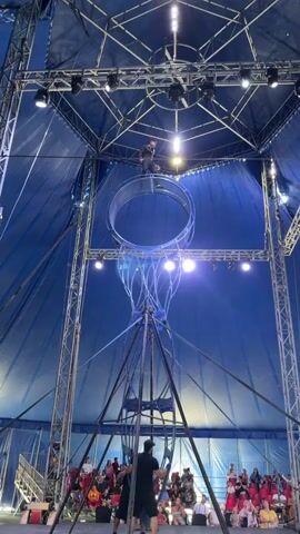 The video shows a circus performer on a large metal apparatus suspended from the ceiling of a blue tent. The apparatus consists of a large ring with a series of curved metal rods extending downwards, resembling a bell shape. The performer, dressed in black, is standing on the ring and appears to be walking and balancing. The performer is illuminated by spotlights. Below the apparatus, an audience is seated in rows. Several other individuals, possibly crew members, are visible on the ground level. The tent structure is visible, with metal beams and rigging supporting the apparatus and lighting. The overall scene depicts a circus performance in progress.