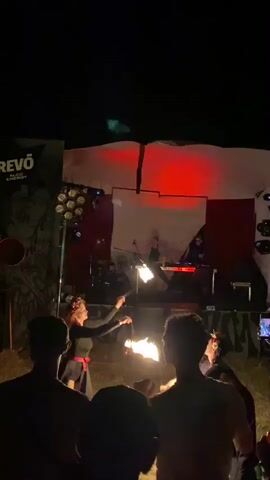 A nighttime outdoor performance is underway. In the foreground, the backs of several spectators are visible, silhouetted against the stage lighting. On the stage, two individuals are performing with fire. One individual, wearing a black top and skirt with a floral headdress, spins flaming objects. Another individual, in a dark outfit and headdress, also manipulates flaming props. Behind them, two DJs are set up at consoles, with audio equipment and lights illuminating the stage. The backdrop consists of a white sheet with red accents, and a banner with text, partially obscured, is visible on the left side. The ground appears to be grass. The overall atmosphere is one of an evening entertainment event.