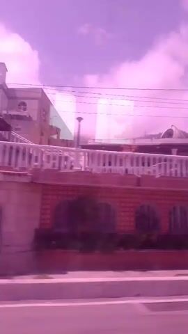 This media file appears to be a short video recording taken from a moving vehicle, likely a bus or car, through a window. The overall scene is dominated by a strong pink or purple hue, suggesting either a filter, post-processing, or unusual lighting conditions.

The primary subjects are buildings and trees, viewed from a street-level perspective. In the first few seconds, a building with a red brick facade and white railings is visible. It has arched windows and a central entrance. Above this, another structure with a lighter facade and what appears to be a rooftop canopy can be seen, set against a hazy sky with clouds. Utility poles and wires are also present.

As the recording progresses, the view shifts to show trees with lush green foliage. Reflections or superimposed images, appearing as green squares, are visible on the window through which the recording is made, obscuring parts of the scene. In one section, a large mural or sign with a sun-like graphic and blue wavy lines is partially visible behind the tree. Further along, glimpses of other buildings and trees suggest a street or roadside environment.

The activity is simply passing by these sights. The time of day is difficult to ascertain due to the color cast, but the presence of clouds and the general brightness might suggest daytime. No people are clearly discernible, and the overall mood is neutral, overshadowed by the visual distortion. The location cues are minimal, but the prompt specifies Macuto, Venezuela, which would place the scene within a coastal or urban environment of that region. No visible text is clearly legible.