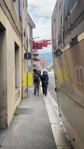 Two people with backpacks walk down a sidewalk next to a street. The sidewalk is made of rectangular gray tiles. There is a beige building to the left and a bus on the right. A bright yellow box stands on the sidewalk. Overhead, there is a cable car infrastructure with a red frame and a white gondola. Cars drive on the street. In the background, buildings line the street, with mountains visible further away under a partly cloudy sky.