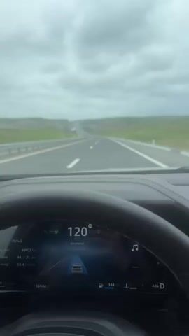 From the driver's perspective, the video shows a car traveling on a highway under an overcast sky. The car is going 120 kilometers per hour. The road is asphalt and appears to be in good condition. The highway is bordered by grassy areas on either side and a metal guardrail on the left.