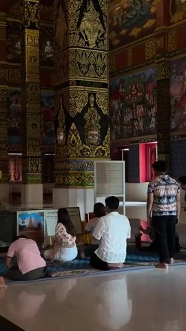 A group of people are sitting on a blue mat on a shiny floor in what appears to be a temple. The people are facing away from the camera. They are dressed in casual clothing. In the background, there are large, ornate pillars decorated with gold and black patterns, as well as murals on the walls. There is also a shrine-like structure to the left.
