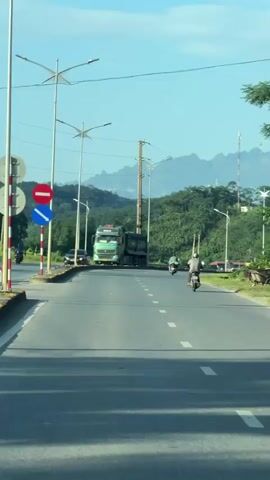 The camera, possibly positioned within a moving vehicle, faces down a straight two-lane road in Vietnam. White dashed lines divide the lanes. On the left side of the road stands a large green dump truck, a small black car, and a sign indicating a no-stopping zone and a mandatory right turn. Utility poles line the side of the road, interspersed with modern streetlights. A man on a motorcycle travels away from the camera in the same direction as the camera. The scenery changes with a light blue delivery truck. In the distance, lush green mountains are visible under a clear blue sky with sparse clouds. The overall atmosphere is bright and sunny.