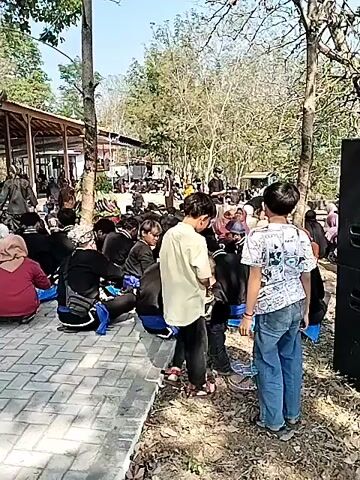 The video shows a gathering of people, many of whom are sitting on the ground on blue mats. They are gathered in an outdoor area with a building in the background that has a traditional style roof. There are trees and a paved area in the foreground. Some people are wearing traditional clothing. There are also children present, with one child in a blue shirt in the center of the frame playing. The overall atmosphere appears to be one of a community event or religious gathering.