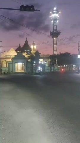 A motorcyclist wearing a red shirt and red cap rides a scooter on a paved road. The scooter's headlight illuminates the road. In the background, a mosque with a tall minaret stands against a twilight sky. The mosque features a dome, several spires, and illuminated windows and architectural details. Traffic lights are visible in the upper left corner of the frame. The scene is captured during dusk or dawn, as indicated by the soft, diffused lighting and the purple and pink hues in the sky.