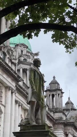 A statue of a man in a suit stands in front of a large building with a green dome. A bird is perched on the statue's head. The statue is covered in moss. The sky is overcast.
