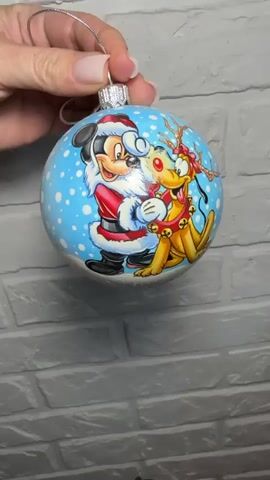 A hand holds a Christmas ornament that is being rotated. The ornament is round and white, with glitter. One side of the ornament has a picture of Mickey Mouse dressed as Santa Claus and Rudolph the reindeer. The other side of the ornament has red lettering that says "Happy New Year" in Russian. The background is a white brick wall.