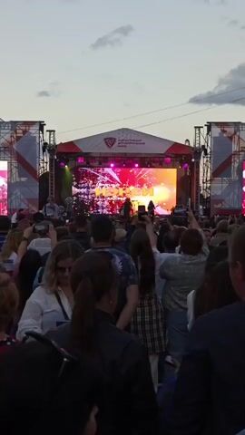 This is a vertical, medium-shot video of a live music concert taking place outdoors at dusk. A large crowd of people is gathered, facing a brightly lit stage. On the stage, a band appears to be performing, with several figures visible in silhouette against the vibrant screen behind them. The screen displays colorful, abstract visuals and text, including what appears to be a band name in large, yellow lettering and smaller text below. Above the stage, a banner is visible with text that includes "ЗЛАТОУСТОВСКИЙ МЕТАЛЛУРГИЧЕСКИЙ ЗАВОД" (Zlatoust Metallurgical Plant), indicating the sponsor or location of the event in Zlatoust, Russia. The sky is a pale blue, suggesting the sun has recently set, and there are a few clouds. Many audience members are holding up their phones to record or take pictures of the performance, reflecting the energetic and engaged atmosphere of the event. Some people in the crowd are smiling and appear to be enjoying the music. The overall mood is festive and lively.