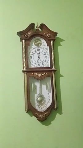 The video shows a close-up shot of a pendulum clock mounted on a light green wall. The clock is made of dark wood with gold-colored accents. It has a white face with black numerals and hands. The pendulum is visible swinging back and forth behind a glass panel. The clock appears to be functioning normally.