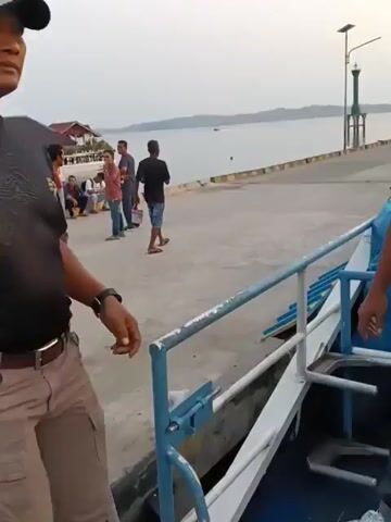 A group of men are gathered on a pier near the water. In the foreground, a man in a black t-shirt and cargo pants walks towards a boat. He has a dark complexion and is wearing a hat. He appears to be a security guard or official, as he has a badge on his shirt with the Indonesian flag. To the right, a boat with blue and white trim is docked. In the background, several other men are standing or sitting on the pier. One man in a red and grey t-shirt is facing away from the camera. Another man in a black t-shirt and blue shorts is looking towards the water and holding a small object. Further in the distance, a small boat is moving across the water, and a hilly, tree-covered landscape is visible on the horizon. The sky is overcast and the lighting suggests it is late afternoon or early morning.