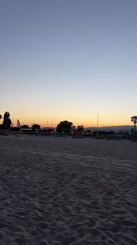 The video shows a sandy beach at dusk. The sand is textured with ripples and appears wet in places. In the background, there are some palm trees and buildings with lights on. The sky is a gradient of blue and orange, indicating sunset. In the foreground, a person's bare foot is visible as they walk across the sand, leaving footprints. The camera angle is low, looking down at the sand and the person's leg.