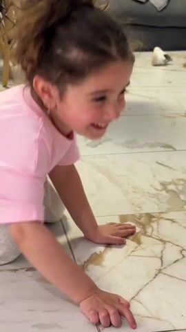 A little girl with brown hair in a messy bun and wearing a pink t-shirt crawls toward a baby in a light blue shirt. Both are on a marbled floor. The girl crawls on her hands and knees, smiling and looking at the baby. The baby looks back at the girl. The girl then sits up, still smiling. In the background, there's furniture including a couch, chairs, and clothing items.