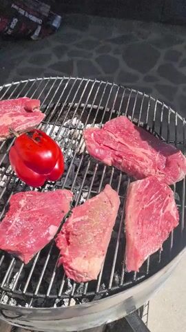 A close-up shot shows several raw steaks and a red bell pepper on a grill. A pair of metal tongs is used to flip one of the steaks. The steaks are seasoned with salt and pepper. The grill is hot, as evidenced by the wisps of smoke rising from the coals. The lighting suggests it is daytime.