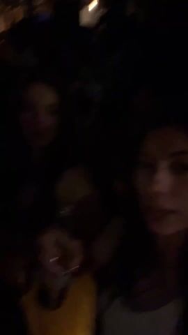 The video takes place at night, likely in a nightclub or bar setting, as suggested by the low light conditions and the presence of multiple people in the background. Two women are the main focus, visible in the foreground. They appear to be smiling and singing along to music. One of the women holds a glass of liquid, suggesting they might be drinking. Other individuals are seen moving around them, including a man in a white shirt. The camera movement is unsteady, indicating it is hand-held, and the overall scene appears lively and social.