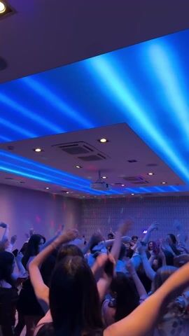 The video shows a group of people dancing in a room with blue ceiling lights and disco lights on the walls. People are raising their arms and jumping, some are recording the scene with their phones. The room has a modern design with a drop ceiling and recessed lighting. There's a projector mounted on the ceiling. The atmosphere is festive and energetic.