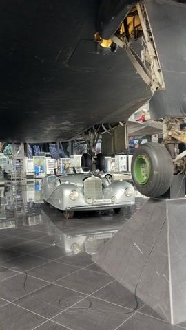 The video shows a classic silver Mercedes-Benz convertible parked beneath the undercarriage of an airplane. The airplane's landing gear, including a large tire with a green rim, is visible. The car is positioned directly in front of the landing gear. The scene appears to be an indoor exhibition or museum, with polished black tiled flooring reflecting the objects above. In the background, there are displays and exhibits suggesting a museum environment. The lighting is bright, illuminating the car and the airplane's components.