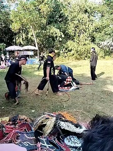 The video shows an outdoor gathering with several people on a grassy area. In the foreground, there are piles of colorful fabric items, possibly costumes or decorations. In the mid-ground, several individuals are engaged in what appears to be a performance or demonstration. One person in the center is wearing black traditional clothing and is holding a circular object with strings attached. Another person, also in black, is on the ground with others around them. Further back, more people are observing. The background consists of trees and some temporary structures like tents or awnings. The overall atmosphere seems to be a cultural event or performance taking place in a park-like setting.