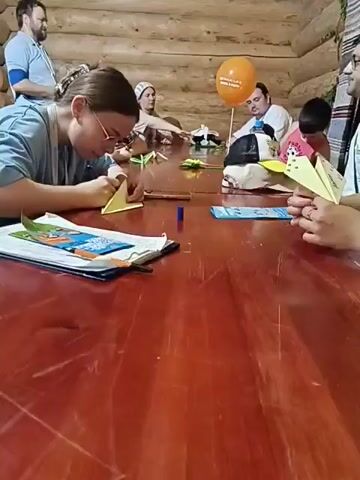 A group of people, including adults and children, are gathered around a large, polished wooden table. The room appears to be rustic, with exposed wooden log walls and ceiling beams. Several people are engaged in what looks like an arts and crafts activity, specifically origami. One woman in the foreground, wearing glasses and a light blue shirt, is intently folding a yellow piece of paper. In front of her is a notebook or binder. Other individuals are also seen folding paper and interacting with materials on the table, which includes colorful paper, markers, and possibly instructions. An orange balloon is visible in the background, attached to a chair. The overall atmosphere suggests a workshop or class.