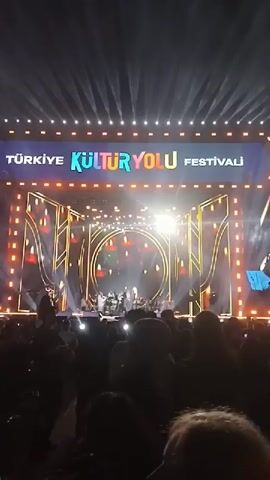 The video shows a live concert at night, filmed from the audience's perspective. The stage is large and ornately decorated with lights and golden geometric patterns. A banner at the top of the stage reads "Türkiye Kültür Yolu Festivali" which transitions to "T.C. Kültür ve Turizm Bakanlığı". A band is performing on the stage, bathed in bright lights. The audience is visible in the foreground, silhouetted against the bright stage lights. Many people are holding up their phones, presumably recording the performance.
