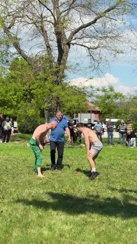 Two shirtless men are about to wrestle in a grassy field under a large tree. A man in a blue shirt and black pants stands between them, presumably as a referee. Several people are watching in the background. The weather appears sunny and mild, with some clouds in the sky.