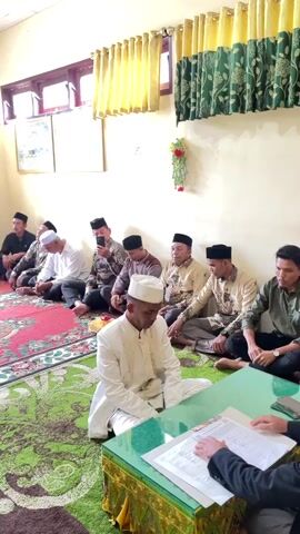 The video shows an indoor gathering of several men seated on the floor, facing towards the viewer. Most of the men are wearing traditional attire, including white or patterned shirts and kufis. In the foreground, a man in a white outfit and kufi is seated and looking down. In front of him, there is a green table with papers and hands visible, suggesting some form of formal activity. The room has yellow curtains and green patterned rugs on the floor. The lighting appears to be natural, coming from windows. The overall atmosphere suggests a religious or ceremonial event, possibly a wedding or a similar gathering.