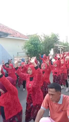 A group of women wearing red outfits and headscarves are dancing in the street. They are raising their arms in unison. A man in an orange shirt is riding a motorcycle in the foreground, facing away from the dancers. There are houses and trees in the background. The sun appears to be setting.