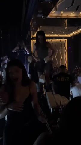 This is a video of a crowded nightclub or performance venue. The lighting is dim and colorful, with purple and white lights illuminating the scene. In the foreground, several people are visible, some dancing and others watching. One woman in the foreground is wearing a black dress. Further back, a stage area is visible with several performers. Four women are on the stage, dressed in revealing outfits, some holding props like a pole and a hat. The stage is lit with purple lights, and there are speakers and lighting equipment visible above. The audience appears to be engaged with the performance, with many people raising their hands. The overall atmosphere is energetic and lively.