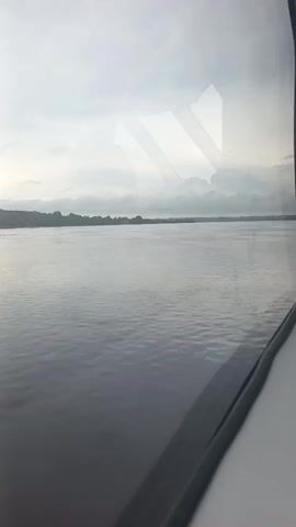 The video shows a wide river with a cloudy sky. The water is calm and appears dark. On the far side of the river, there is a distant treeline. The view is through a window, and there are reflections on the glass, including a diagonal pattern of light. The camera is stationary, capturing a static view of the river and sky.