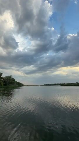 A video shows a body of water with a shoreline of trees and foliage in the distance. The sky is partly cloudy with patches of blue sky visible. Sunlight is breaking through the clouds in some areas, creating rays of light. The water is calm with gentle ripples. The camera is positioned low, likely from a boat or close to the water's surface. The time code indicates the video is only a few seconds long, capturing a tranquil natural scene.