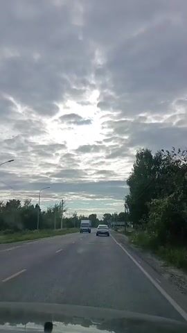The video shows a view from a vehicle driving down a paved road. The sky is overcast with patchy clouds, and the sun is visible peeking through. On either side of the road, there are grassy areas with trees and some buildings. In the distance, other vehicles, including a white van and a white car, can be seen on the road. The vehicle appears to be driving through a rural or suburban area. There are power lines overhead, and a lamppost is visible at one point. As the video progresses, the view shifts to show more of the roadside, including some construction materials like piles of gravel or sand, and some modular buildings. A Russian flag is visible on a flagpole in the distance. The scene suggests the vehicle is continuing along a road with development and infrastructure present.