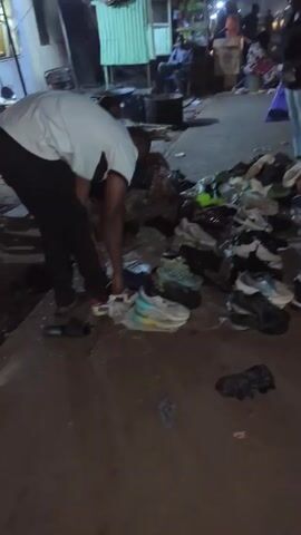 The video depicts an outdoor scene at night, likely a street market. The main focus is a pile of various shoes displayed on the ground. A person wearing black pants and a light-colored shirt is bending over, seemingly looking at the shoes. Another individual in light-colored pants and a dark top is standing nearby. In the background, people are walking along the road, and there are various vehicles, including auto rickshaws (Tuk-Tuks), parked. Streetlights illuminate the area. The general atmosphere suggests a bustling night market environment.