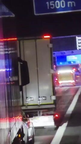 A nighttime, first-person perspective video shows a highway scene. The camera is positioned behind a large white semi-truck with red lights on its trailer. Ahead, on the right side of the highway, are multiple emergency vehicles with flashing lights, including what appears to be a fire truck and an ambulance. A large blue highway sign is visible above the emergency vehicles, indicating destinations such as München, Würzburg, and Fulda, with distances and highway numbers. Traffic appears to be stopped or moving very slowly.