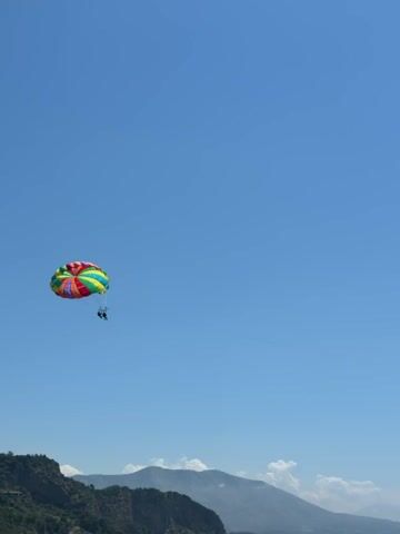 A colorful parachute carrying two people is seen soaring through a clear blue sky over a mountainous landscape. The parachute is multicolored, with stripes of red, orange, yellow, green, and blue. Two figures are visible suspended beneath the parachute, appearing to be enjoying a tandem parasailing experience. The background features rolling hills and mountains, suggesting an outdoor recreational setting. The video appears to be filmed from a slightly elevated position, looking upwards towards the sky.